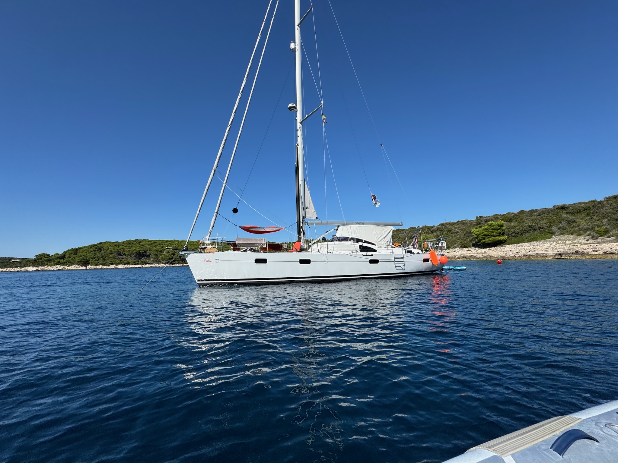SV Nellie at anchor in a calm cove, deep blue water with a green headland behind